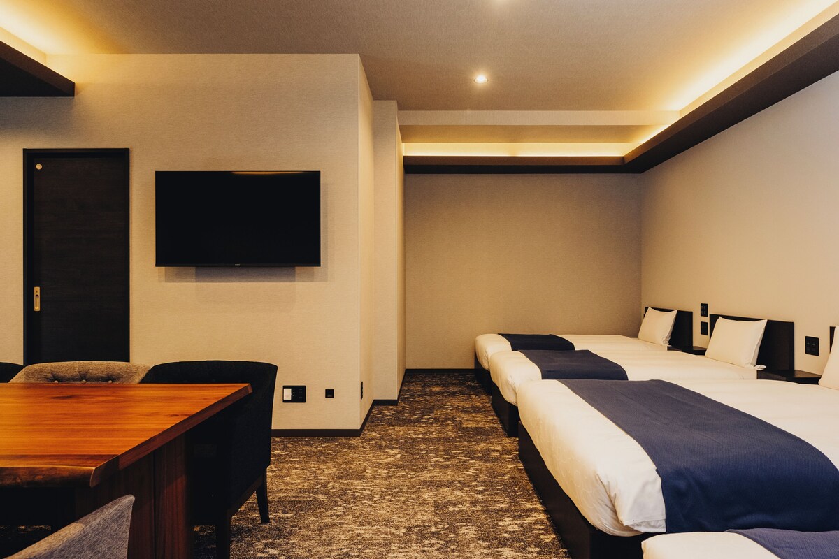 Een hotelkamer met een houten eettafel, twee bedden met donkerblauwe beddengoed, een wandgemonteerde flatscreen televisie en een vloerbedekking.