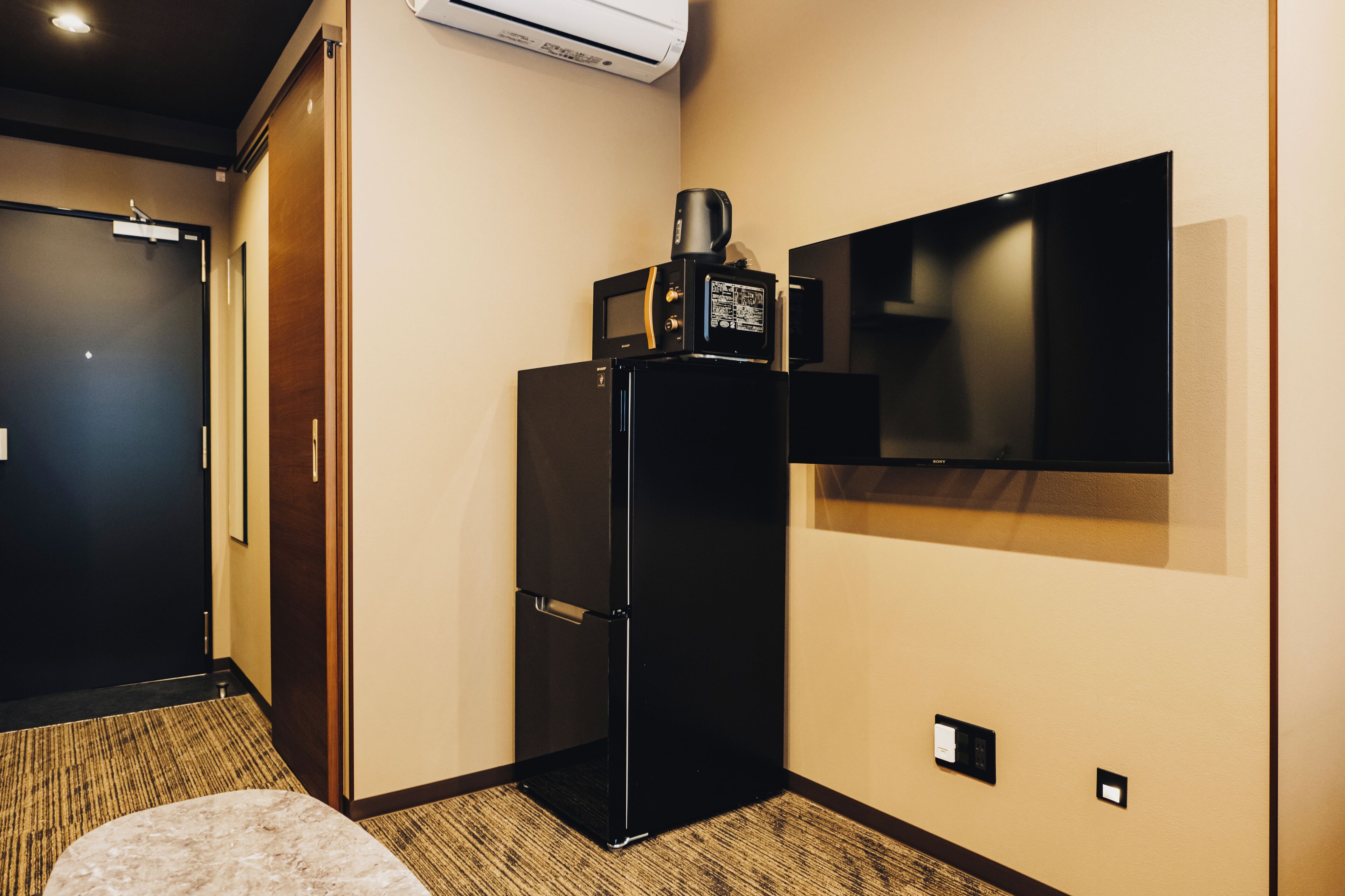 A hotel room with a black refrigerator, a flat-screen TV mounted on the wall, a carpeted floor, and a door with a handle.