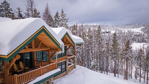 Una cabaña de troncos con cimientos de piedra, terraza de madera y techo cubierto de nieve.
