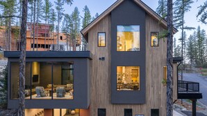 A modern two-story house with large windows and a balcony, surrounded by trees.