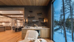 A modern kitchen with a stone backsplash, wooden cabinets, and a large window offering a view of a snowy forest.
