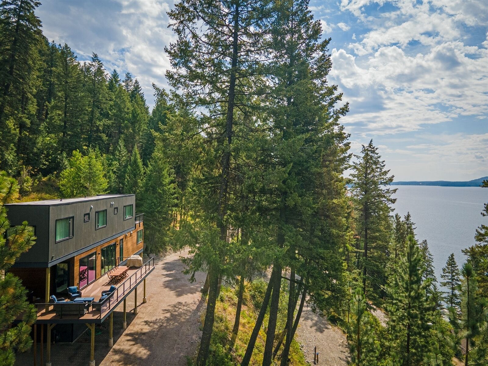 Una cabaña moderna con terraza y asientos al aire libre, rodeada de un bosque denso y con vistas a un lago.