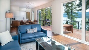 A modern living room with a blue sofa, a glass coffee table, and a view of a deck with outdoor furniture. - NEW private hot tub & amazing lake views (Whitefish)