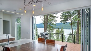 A modern kitchen with a large island, a dining area with a wooden table and chairs, and a view of a lake with trees and mountains. - NEW private hot tub & amazing lake views (Whitefish)