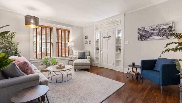 Un salón con un sofá gris, un sillón azul, una mesa de centro redonda y otro sillón blanco. Hay un ventanal con persianas de madera, una puerta blanca con un panel de vidrio y un estante en la pared con plantas.