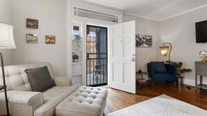 Un salón con sofá, reposapiés y una mesa de centro. Hay una puerta que da a un balcón con vistas a edificios.