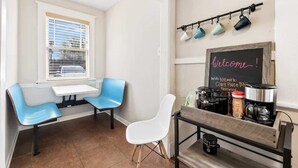 Un café pequeño y acogedor con dos sillas azules, una silla blanca, una mesa y un cartel de pizarra con la leyenda '¡bienvenidos!'.