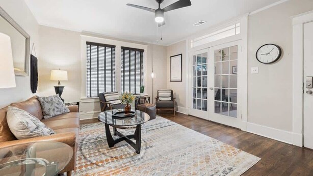 Un salon spacieux avec un canapé en cuir marron, une table basse en verre et un tapis à motifs. On y trouve un ventilateur de plafond, une horloge au mur et une porte donnant sur un balcon.