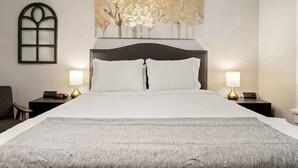 Un lit bien fait, avec une tête de lit sombre, deux tables de chevet avec des lampes, une fenêtre fixée au mur et une grande peinture abstraite au-dessus du lit.