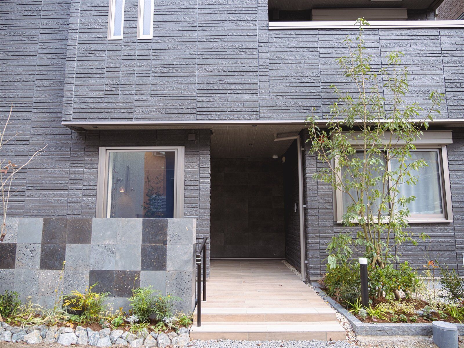 A modern building with a stone facade, a wooden entrance, and a small garden area with plants and a tree.
