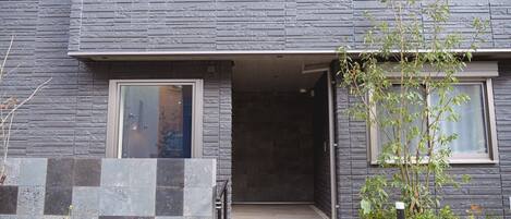 A modern building with a stone facade, a wooden entrance, and a small garden area with plants and a tree.