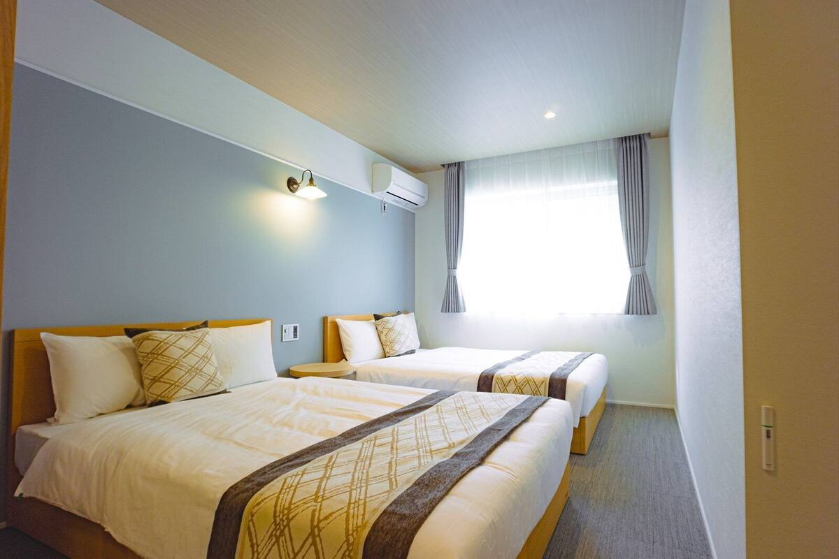 Ett hotellrum med två sängar, ett fönster med gardiner och en väggmonterad luftkonditioneringsanläggning.