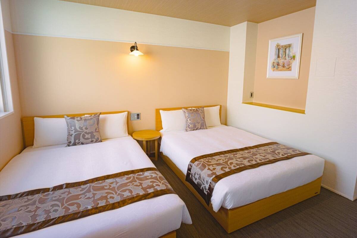 Ett hotellrum med två sängar, var och en med en sänggavel i trä och vitt sänglinne. Mellan sängarna står ett litet runt bord, och ovanför bordet sitter en vägglampa.