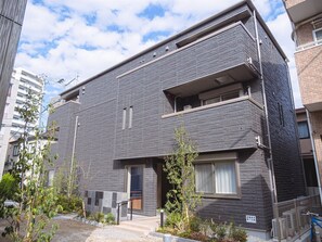 A modern apartment building with a grey facade, multiple balconies, and a small garden area with plants and a bicycle. - GRANDBASESUMIYOSHI (Fukuoka)