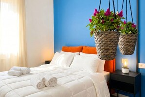 A neatly made bed with white bedding, two hanging woven baskets with plants, a nightstand with a lamp, and a blue accent wall.
