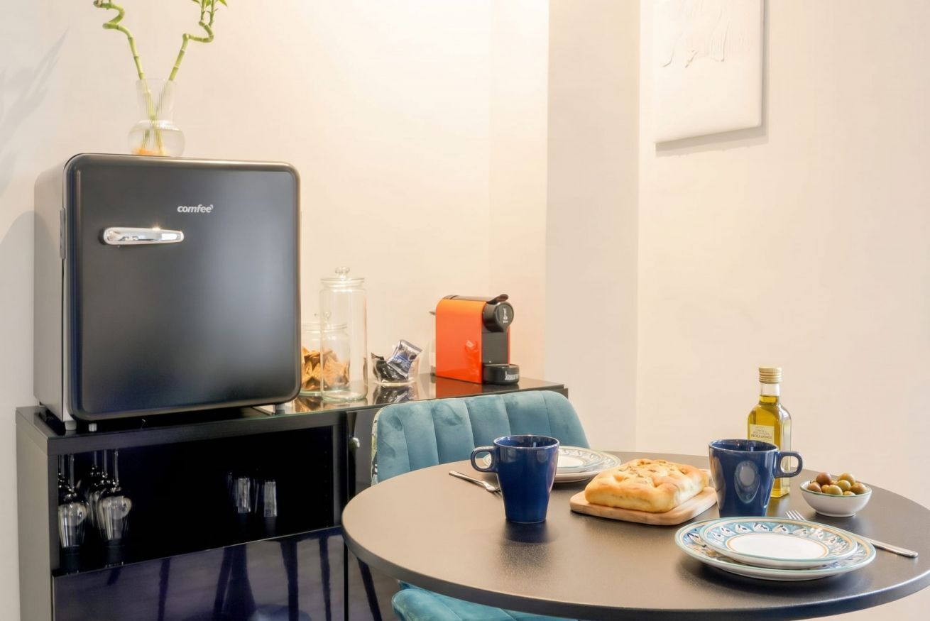 Una cucina compatta con macchina da caffè, piccolo frigorifero, tavolo da pranzo e una sedia blu, e un vaso con piante verdi.