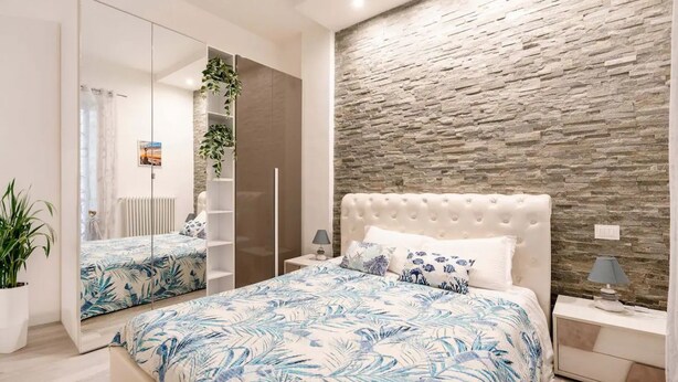 Ein Schlafzimmer mit einem raumhohen Spiegel, einer Wand mit Steinoptik und einem Bett mit floralem Bettbezug.