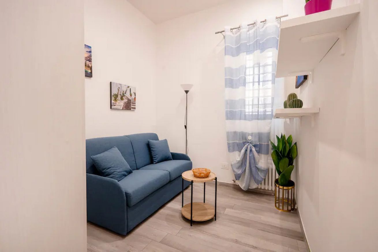 A modern living room with a blue sofa, a small round table, a floor lamp, a window with a curtain, and a shelf with a cactus.