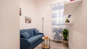 Ein modernes Wohnzimmer mit einem blauen Sofa, einem kleinen runden Tisch, einer Stehlampe, einem Fenster mit Vorhang und einem Regal mit einem Kaktus.