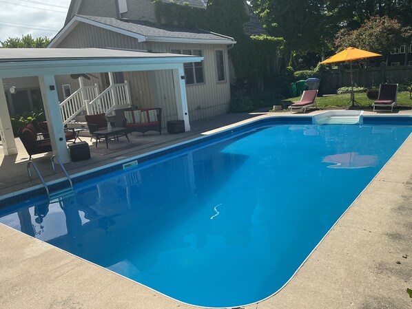 A rectangular swimming pool with a white deck, a covered seating area, and a wooden fence in the background.