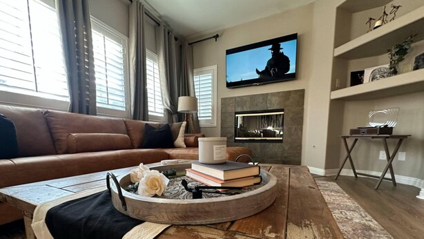 A living room with a brown leather sofa, a coffee table with books and flowers, a fireplace, and a television on the wall. - Spacious 4-bedroom townhome with Pool, WiFi, AC in marvelous Murrieta (Murrieta)