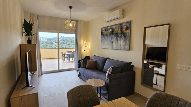 Un salon comprenant un canapé, un téléviseur, une petite table et un balcon donnant sur un espace verdoyant.