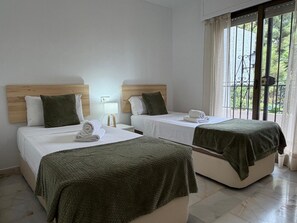 A hotel room with two beds, each with a wooden headboard, white linens, and a green blanket. There is a window with curtains, and a small table with a lamp.