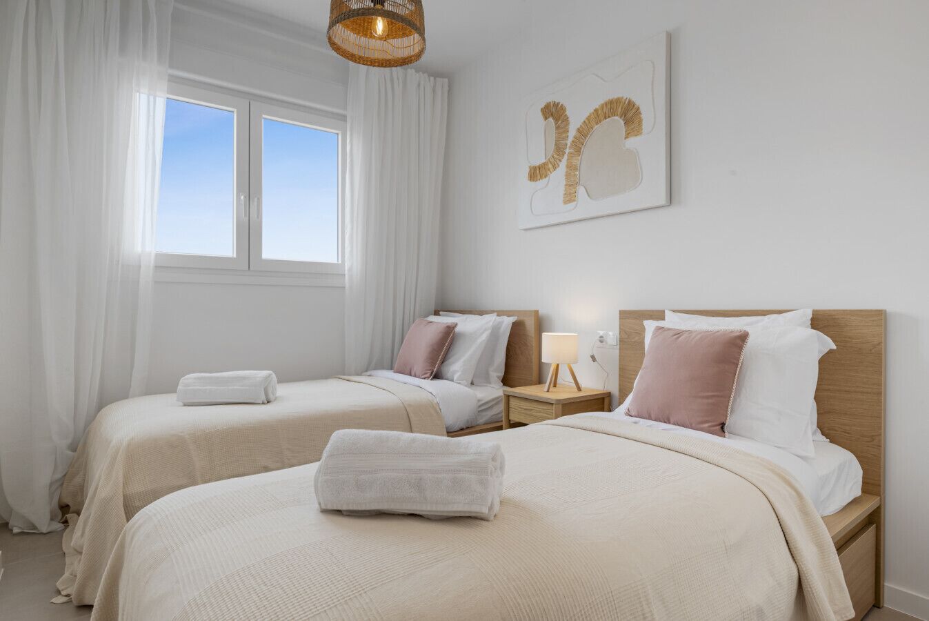 A hotel room with two beds, white linens, a wooden headboard, a bedside table with a lamp, and a window with sheer curtains.