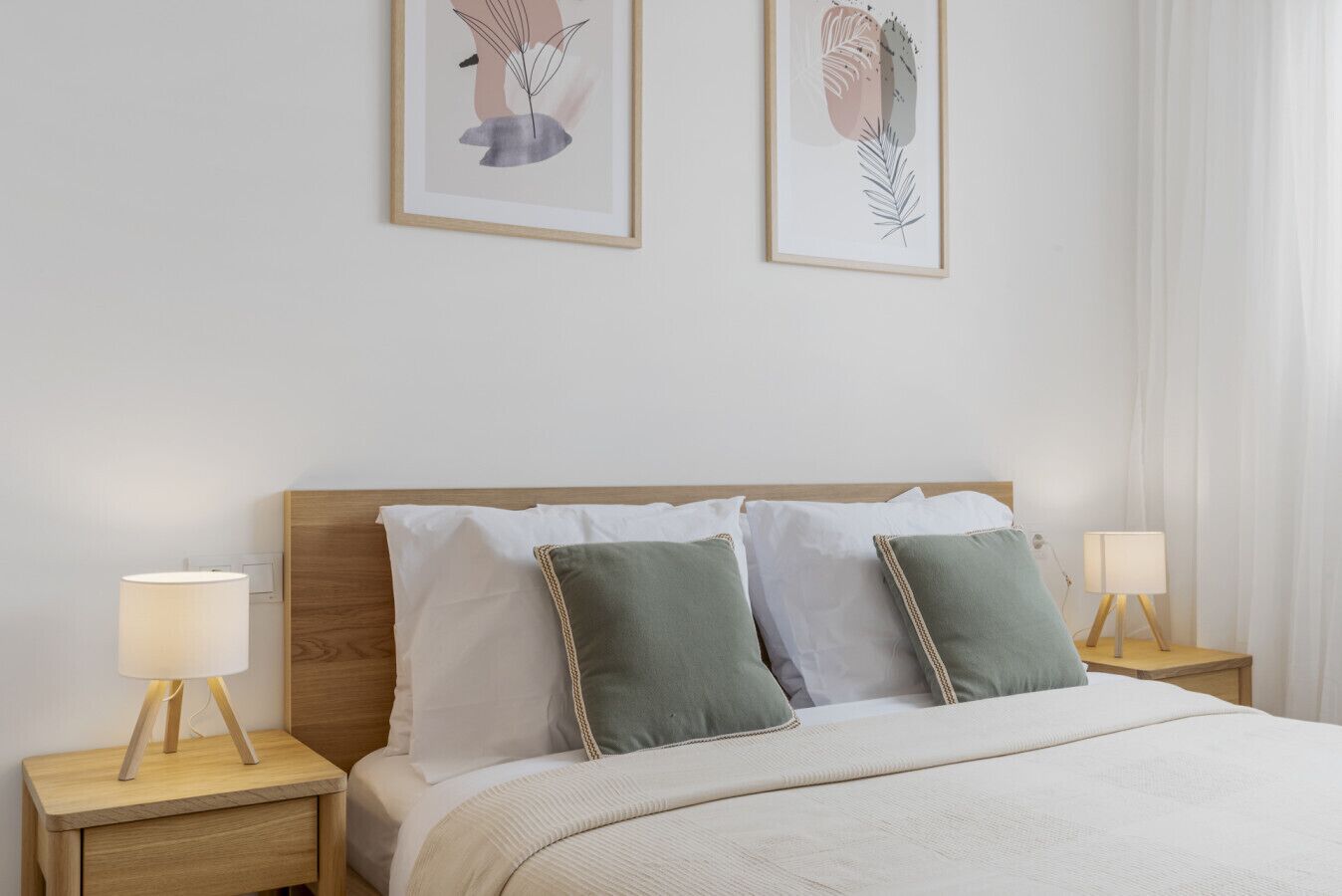 A neatly made bed with white and green pillows, two bedside lamps, and two framed botanical prints on the wall.