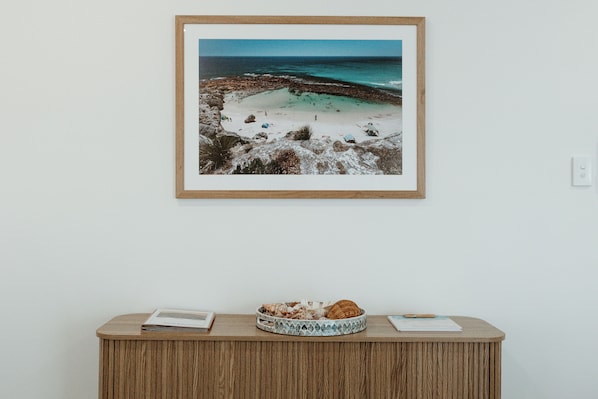 A framed beach scene with a wooden side table holding a book, a tray with bread, and a notebook. - Sunset Hideaway - In Kingscote (Kingscote)