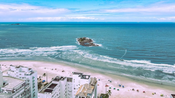 A coastal hotel with multiple floors, a beach view, and a rocky outcrop in the sea. - Apto na Praia - Ar condicionado e Churrasqueira (Guaruja)