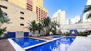A rooftop pool with a wooden deck, surrounded by palm trees and a cityscape in the background. - Apto na Praia - Ar condicionado e Churrasqueira (Guaruja)