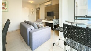 A modern living room with a grey sofa, a dining table with chairs, a TV mounted on the wall, and a window with a view. - Apto na Praia - Ar condicionado e Churrasqueira (Guaruja)