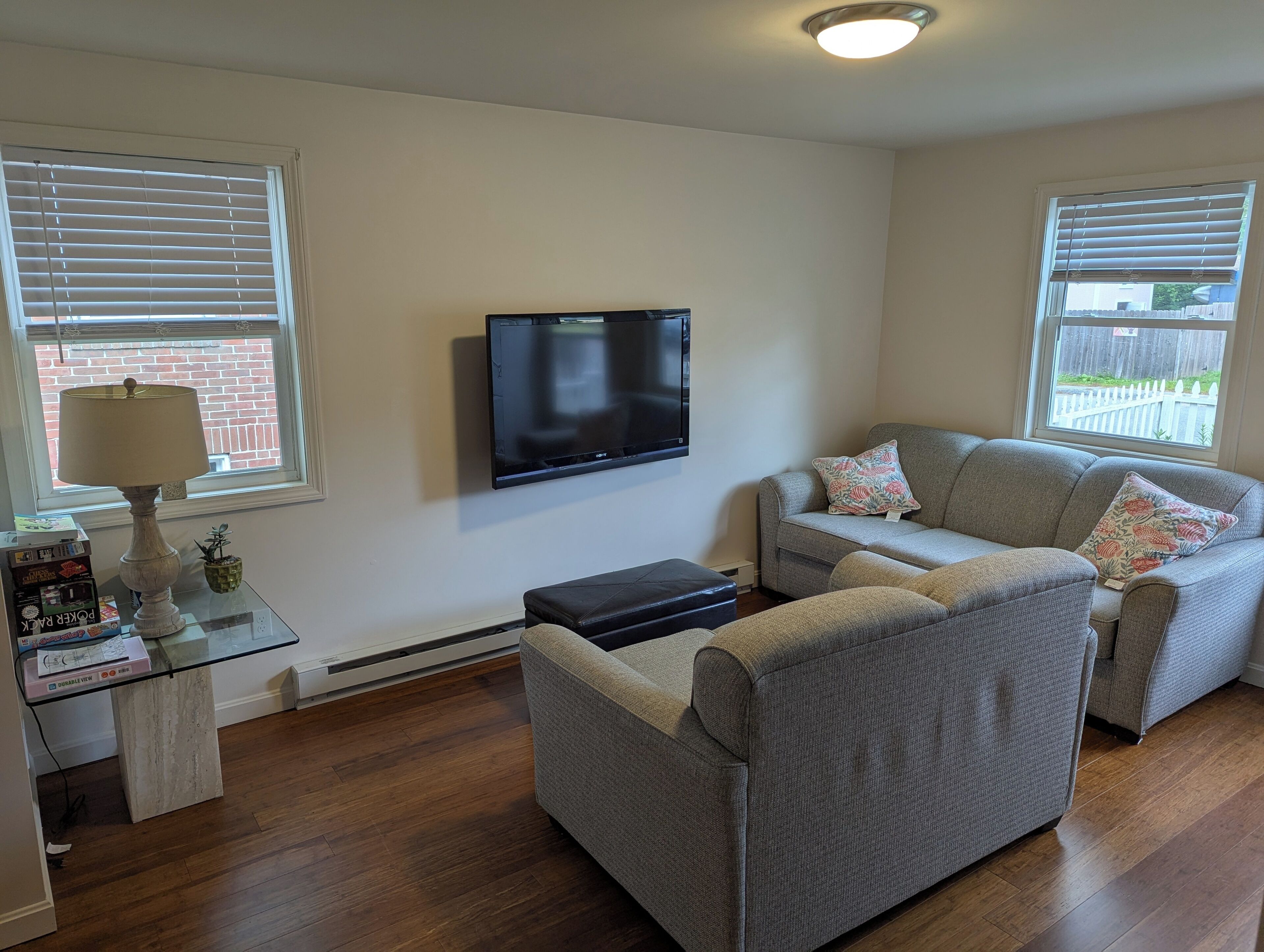 A living room with a flat-screen TV, a grey sofa, a small table with a lamp, and a window with blinds.