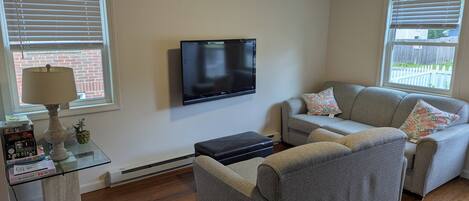 A living room with a flat-screen TV, a grey sofa, a small table with a lamp, and a window with blinds.