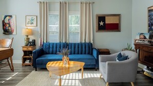 Un salon avec un canapé bleu, une table basse en bois, un fauteuil gris, une table d’appoint en bois avec une lampe, et un mur orné d’un drapeau et d’œuvres d’art.