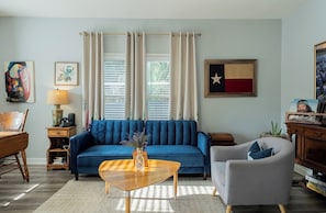 Een woonkamer met een blauwe bank, een houten salontafel, een grijze fauteuil, een houten bijzettafeltje met een lamp, en een muur met een vlag en kunstwerken.