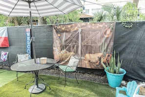 Un patio con una mesa y sillas de metal, un parasol a rayas y un fondo decorativo con cactus y una guitarra.