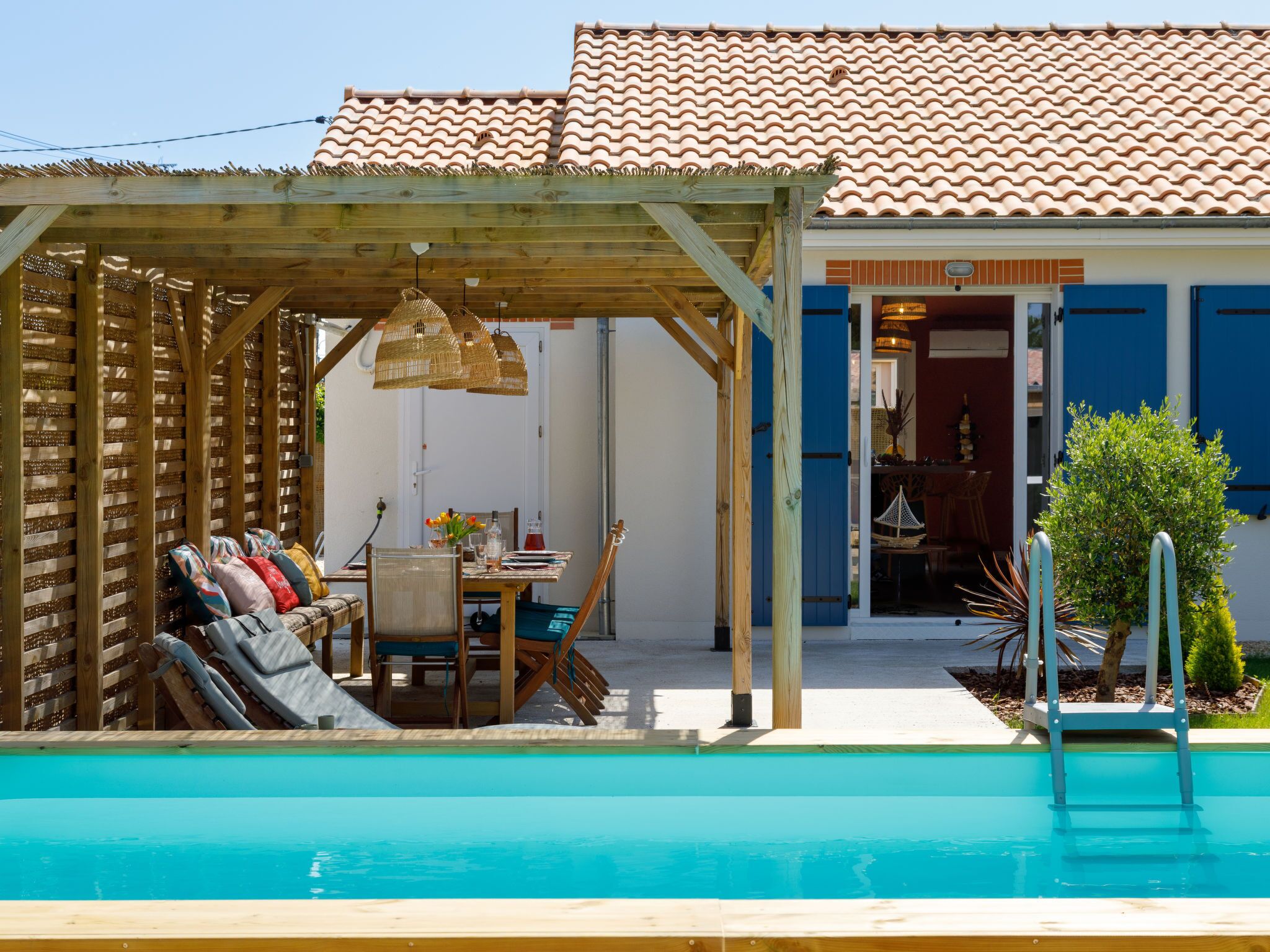 Un espace piscine agrémenté d’une pergola en bois, d’une table à manger avec des chaises, et offrant une vue sur un bâtiment aux portes bleues.