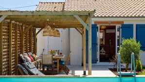 A pool area with a wooden pergola, a dining table with chairs, and a view of a building with blue doors. - La Perle de Jade by Interhome (Pornic)