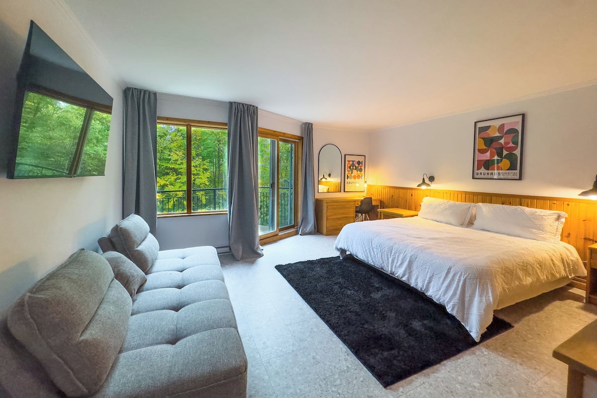 A modern bedroom with a large bed, a sofa, a desk, and a window with a view of trees.