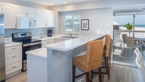Cocina moderna con armarios blancos, microondas integrado y una isla grande con pileta. Hay sillas de mimbre y se ve la playa a través de una puerta de vidrio.