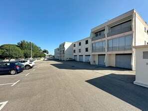 Un parking avec plusieurs voitures, un immeuble à étages doté de balcons et un ciel bleu dégagé.