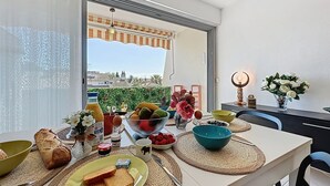 Una mesa de comedor dispuesta con alimentos para el desayuno, como pan, fruta y salsas, en una habitación luminosa con vistas a edificios y un balcón.