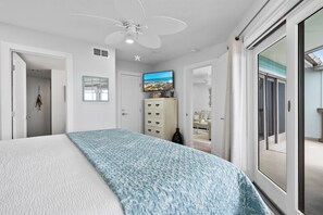 Une chambre à coucher comprenant un lit, un ventilateur de plafond, un téléviseur et une porte-fenêtre coulissante donnant sur un balcon.