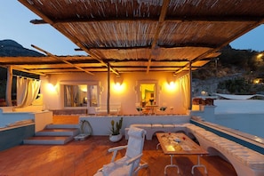 A covered outdoor seating area with a pool, white furniture, and warm lighting.