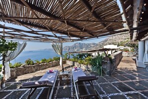 Terraço com duas espreguiçadeiras, uma mesa e vista para o mar e montanhas.
