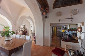 Uma área de refeições com mesa de madeira e cadeiras brancas, uma cozinha com panelas de cobre e vista para outro cômodo com parede amarela.