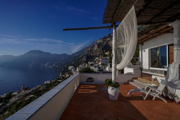 A terrace with a view of the sea and mountains, white outdoor furniture, and a hanging white cloth.