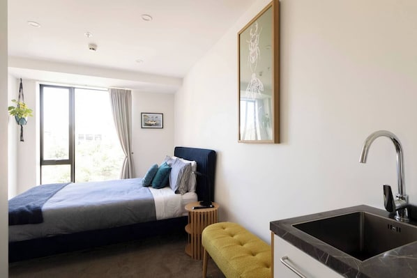 A modern bedroom with a bed, bedside table, bench, and a small kitchenette with a sink.